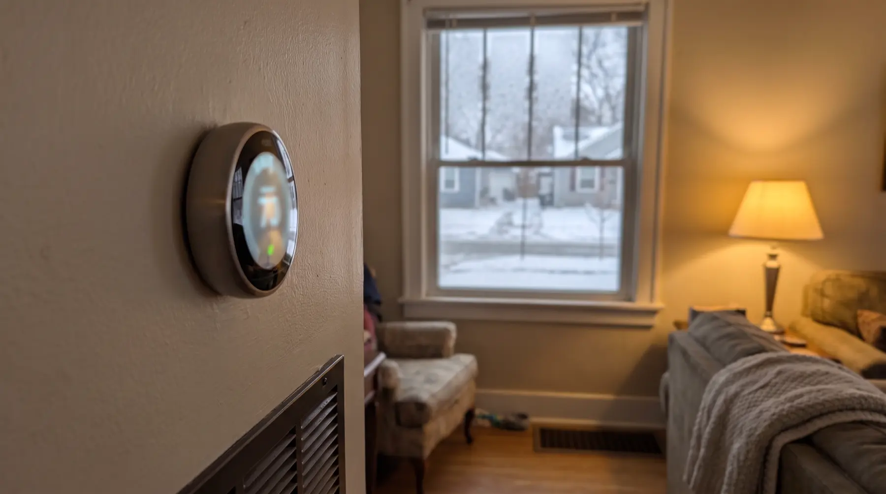 A round smart thermostat on a living room wall with snow outside the window